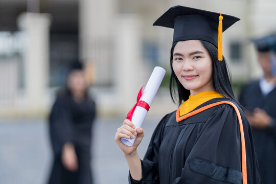 A young beautiful Asian woman university graduate in graduation gown and mortarboard holds a degree certificate stands in front of the university building after participating in college commencement