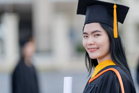 A young beautiful Asian woman university graduate in graduation gown and mortarboard holds a degree certificate stands in front of the university building after participating in college commencement