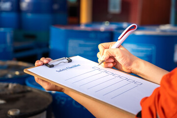 Safety inspection on the checklist document action with blurred background of chemical storage area at factory. Industrial safety concept photo. Selective focus at human's hand part.