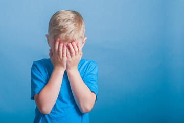 The emotion of surprise. A boy in a blue T-shirt covered his face with his hands against a blue...