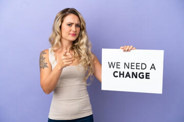 Young Brazilian woman isolated on purple background holding a placard with text We Need a Change and pointing to the front