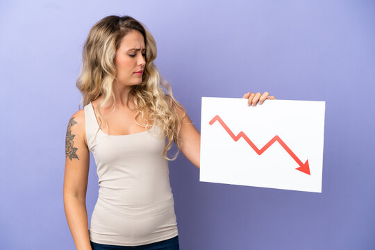 Young Brazilian woman isolated on purple background holding a sign with a decreasing statistics arrow symbol with sad expression