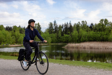 Obraz premium Adult Caucasian Woman Riding a Bicycle on a path by a lake in a modern city park. Spring Evening. Taken in Green Timbers Urban Forest, Surrey, Vancouver, British Columbia, Canada.