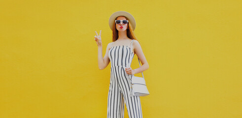 Stylish model woman wearing a summer round straw hat, white striped jumpsuit posing on an yellow background