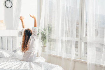  Woman in white bathrobe sitting near the big white window while stretching on bed after waking up.