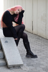 Girl with long pink hair sits on the steps holding her head with one hand and looks forward