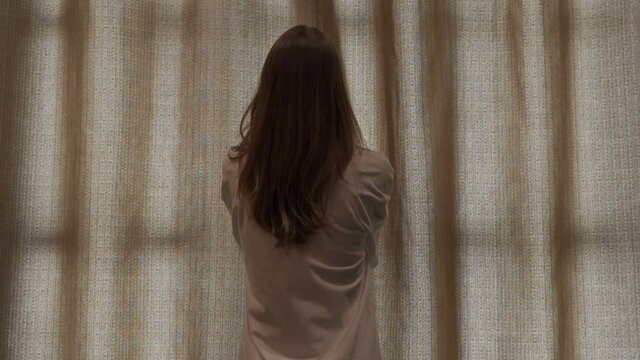 Rear view - a young, fit woman in a white T-shirt opens the bedroom window curtain.