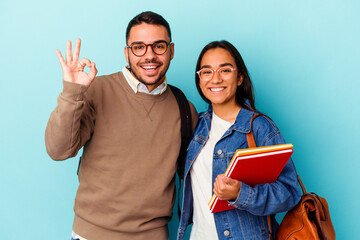 Young mixed race student couple isolated on blue background cheerful and confident showing ok...