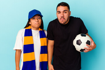 Young mixed race couple cleaning home isolated on blue background shrugs shoulders and open eyes...