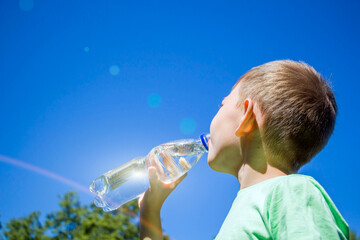 Little boy 6-7 years drinking pure clear water from plastic bottle  against blue sky. Thirsty kid...