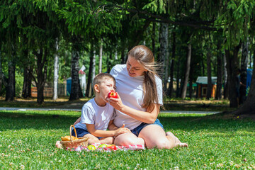 Naklejka na ściany i meble Mom and son eats apple sitting on green grass. Outdoors picnic on summer sunny day in park.