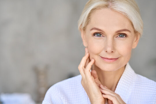 Closeup of happy smiling beautiful senior adult woman of 50s wearing bathrobe at spa hotel looking at camera touching face. Antiage spa procedures advertising. Skin care products concept. Copy space.