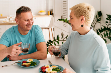 Happy mature man and woman enjoys healthy breakfast together on the table. Looking at each other. Happy retirement life of elderly couple