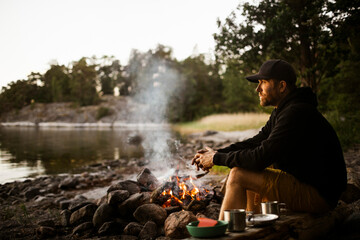 Side view of thoughtful man looking away while sitting by campfire at lakeshore
