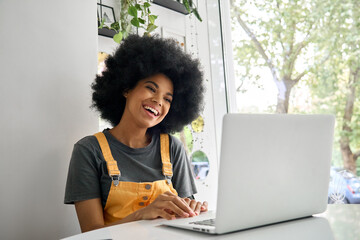 Young happy smiling black teenage girl of 20s with afro hair sitting in cafe indoors speaking with friends on video call using laptop having online web videoconference.