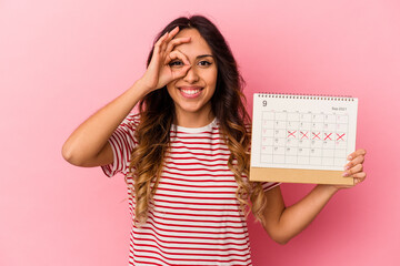Young mexican woman holding a calendar isolated on pink background excited keeping ok gesture on...