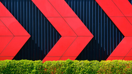 Front view of red arrows pattern on black corrugated metal wall behind green bush fence in exterior...
