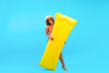 Full length of young black lady holding inflatable lilo and smiling at camera on blue studio...