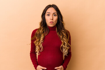Young mexican pregnant woman isolated on beige background shrugs shoulders and open eyes confused.