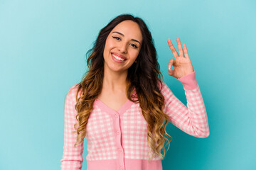 Young mexican woman isolated on blue background winks an eye and holds an okay gesture with hand.