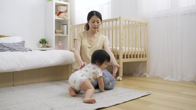 curious asian young child is crawling away after playing with the rug on bedroom floor and ignoring her mother as she is extending arms to offer a hug.