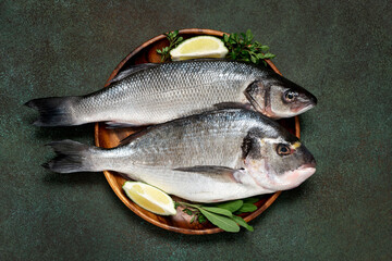 Uncooked fish with lime and herbs in a wooden bowl on a dark green background top view. Raw dorada...