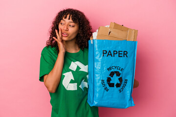 Young caucasian woman holding a recycled plastic isolated on pink background is saying a secret hot braking news and looking aside