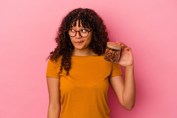 Young mixed race woman holding an almond jar isolated on pink background confused, feels doubtful and unsure.