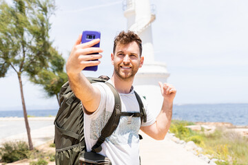 Young Hiker Boy Taking a Selfie in Front of a Lighthouse on the Beach While Smiling