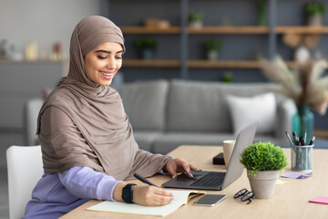 Muslim woman sitting at desk, using computer writing in notebook