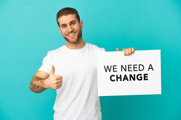 Handsome blonde man over isolated blue background holding a placard with text We Need a Change with thumb up