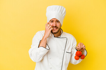 Young caucasian chef man holding tomatoes isolated on yellow background biting fingernails, nervous...