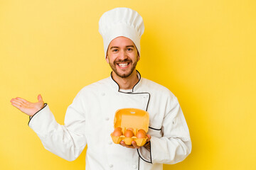 Young caucasian chef man holding eggs isolated on yellow background showing a copy space on a palm and holding another hand on waist.