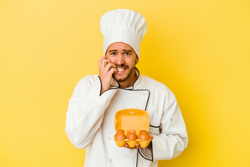 Young caucasian chef man holding eggs isolated on yellow background biting fingernails, nervous and very anxious.