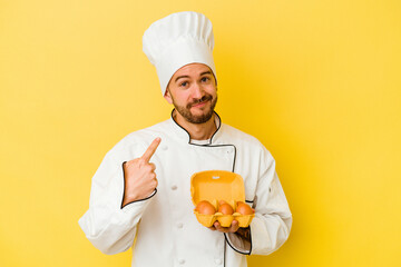 Young caucasian chef man holding eggs isolated on yellow background pointing with finger at you as if inviting come closer.