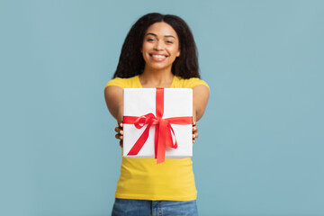 This present is for you. Happy black lady holding wrapped gift box and giving it to camera, standing over blue background