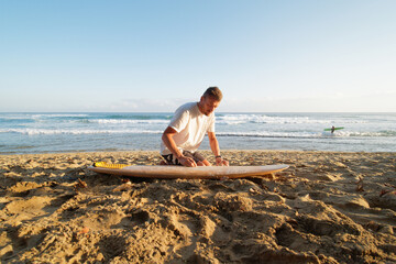 Man on the surf beach waxing surfboard, against ocean with warm tropical water. Dominican Republic.