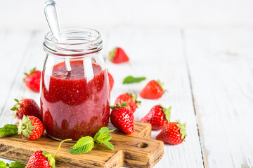 Strawberry jam in the glass jar at white table with fresh strawberries.