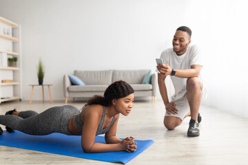 Obraz premium Millennial black couple training as team at home, athletic woman standing in plank pose, her boyfriend noting time