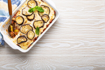 Fototapeta na wymiar Greek mediterranean dish Moussaka with baked eggplants, ground beef in white ceramic casserole with napkin on wooden white background from above, traditional dish of Greece, space for text