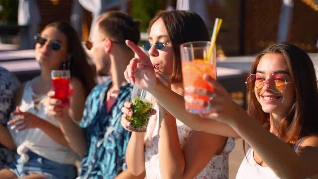 Friends having fun at poolside party clinking glasses with colorful cocktails sitting by swimming pool on sunny summer day. People toast drinking beverages with their feet in a water at holiday villa.