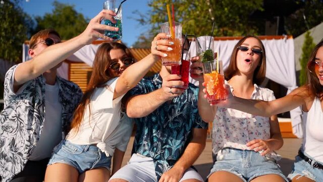 Group of friends having fun at poolside party clinking glasses with fresh cocktails sitting by swimming pool and dancing. People toast drinking beverages at luxury villa on tropical vacation. Slomo.