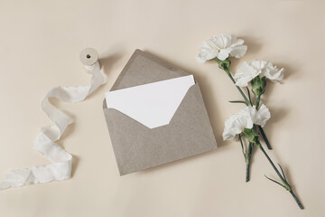 Feminine wedding, birthday stationery composition. Blank greeting card, invitation mockup with craft envelope. White carnation flowers and silk ribbon on beige table background. Flat lay, top view.