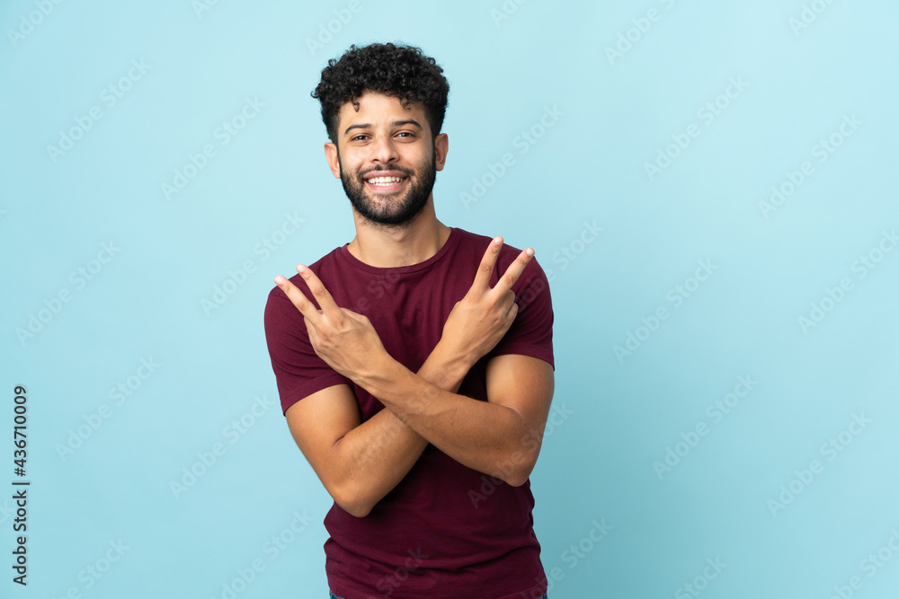 Wall mural young moroccan man isolated on blue background smiling and showing victory sign