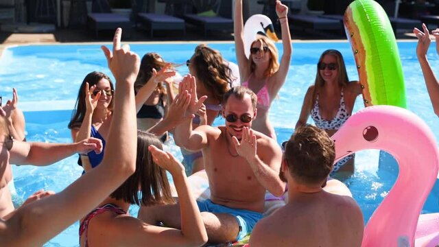 Friends have party in a private villa swimming pool. Happy young people in swimwear dancing, bonding and clubbing with floaties and inflatable mattress in luxury resort on sunny day. Slow motion.