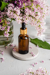 Fototapeta na wymiar Glass brown serum bottle with a pipette for skin care with branches and flowers of lilac on a gray background. Health and beauty concept
