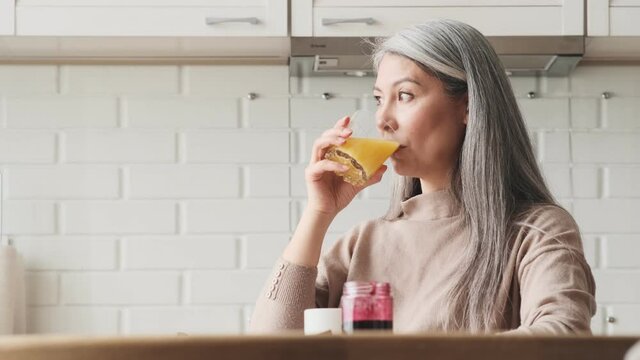 A happy elder woman is drinking a glass of orange juice sitting in the kitchen at home