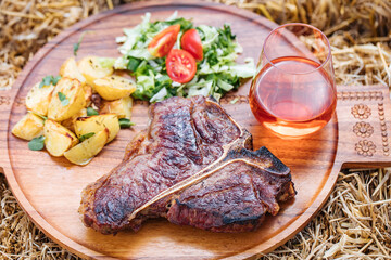 T bone steak served on a wooden board placed on a straw, with salad, potatoes and glass of rose