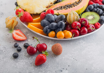 Fresh raw organic summer berries and exotic fruits in white plate on light background. Pineapple, papaya, grapes, nectarine, orange, apricot, kiwi, pear, lychees, cherry and physalis. Macro