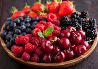 Fresh organic summer berries mix in round wooden tray on dark wooden table background. Raspberries, strawberries, blueberries, blackberries and cherries. Macro. Close up